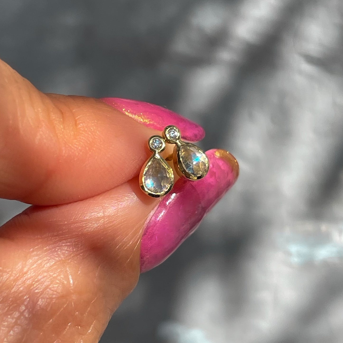 Moonstone Earrings by NIXIN Jewelry held in front of a grey backdrop. Made in 14k gold and bezel set with tiny diamonds above the moonstones.