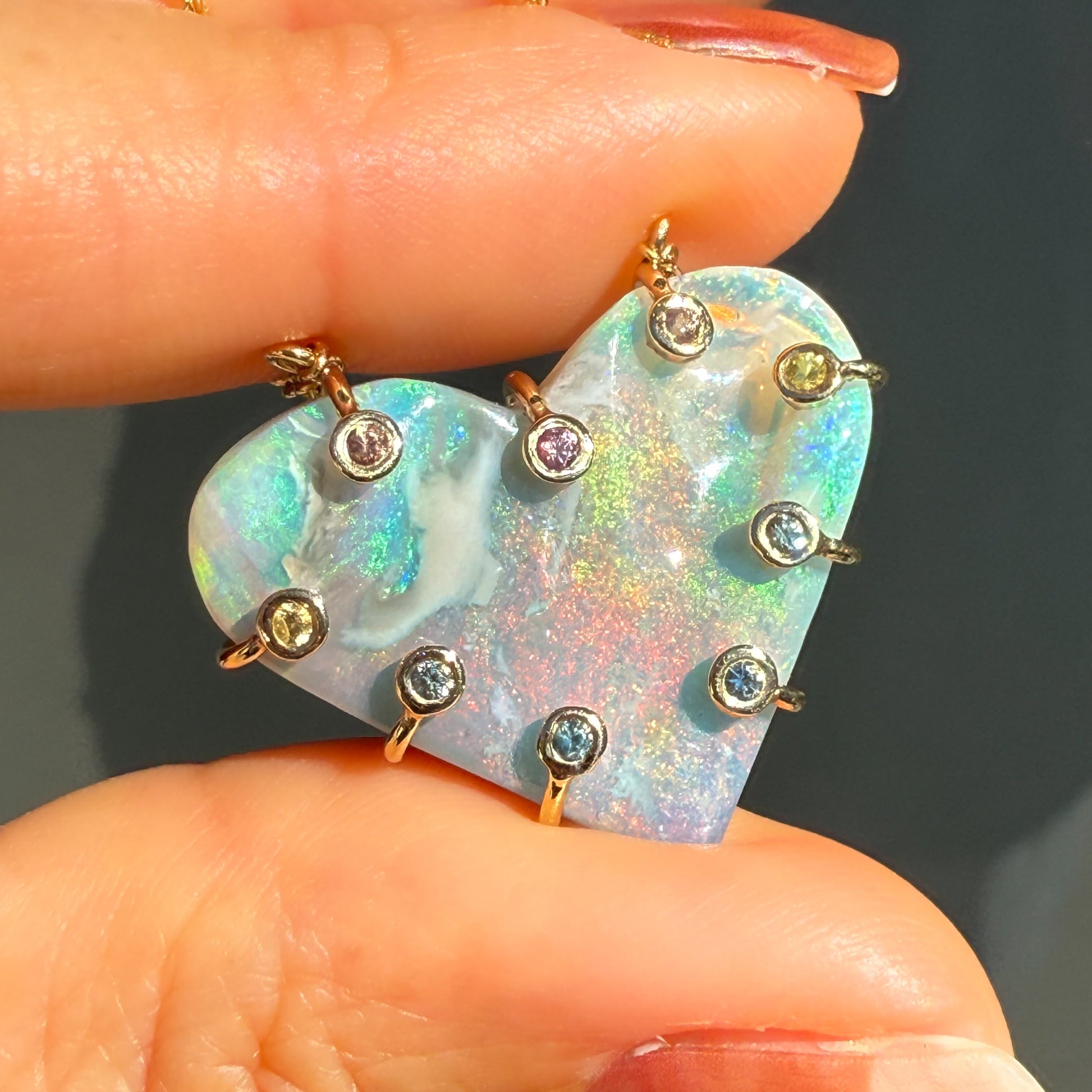 An Australian Opal Necklace by NIXIN Jewelry held between two fingers, emphasizing the vivid play-of-color in the semi black opal. The gold opal necklace features pink sapphire and blue sapphire accents in a bezel setting. This rare opal necklace provides high-contrast luxury.