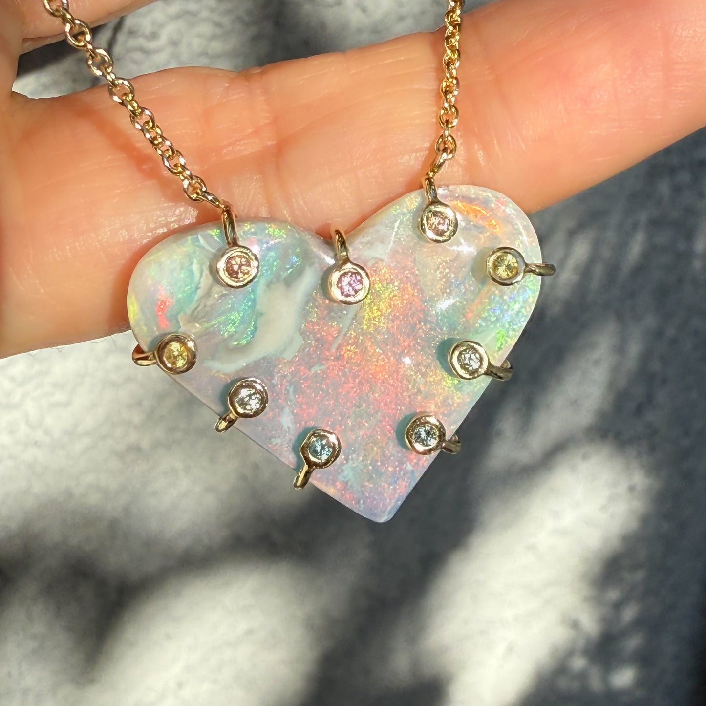 An Australian Opal Necklace by NIXIN Jewelry in bright light, emphasizing the vibrant opal fire across the opal stone. The gold opal necklace is secured by ten sapphires in bezel setting fusing a mix of opal and sapphire. This rare opal jewelry offers vivid color range.