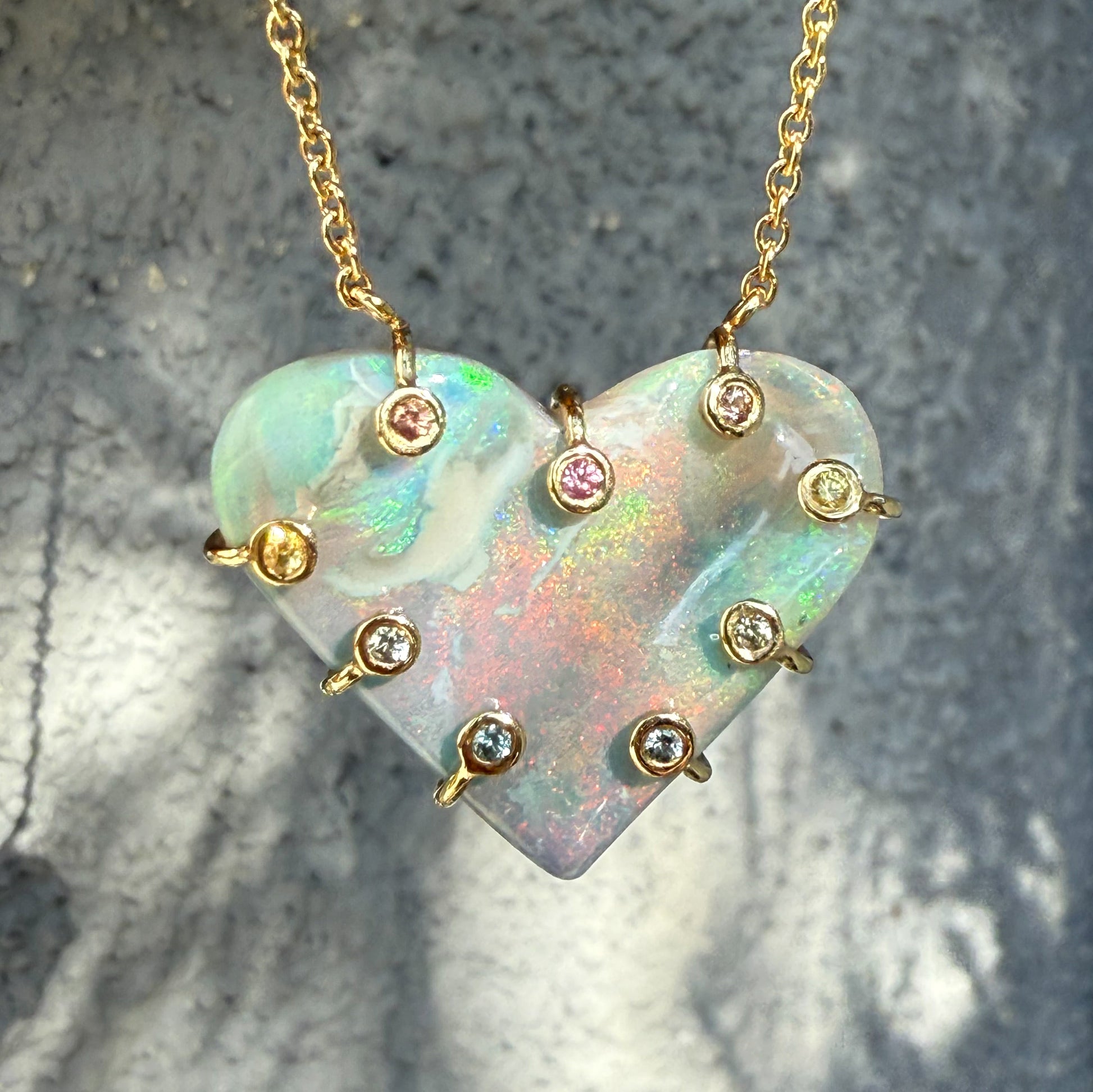 An Australian Opal Necklace by NIXIN Jewelry featuring a shimmering heart-shaped opal pendant against a dark background. The unique opal jewelry design includes a pink sapphire and yellow sapphire among other sapphires accenting the perimeter. This luxury necklace showcases the colorful lightning ridge opal.