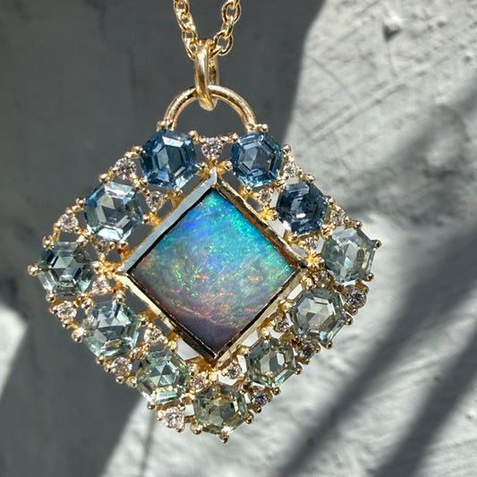 A macro view highlights an Australian Opal Pendant by NIXIN Jewelry. This piece of unique fine jewelry showcases a natural opal center stone displaying vivid neon blue and green hues. The central black opal is framed by a geometric ombré jewelry border composed of hexagon sapphire jewelry prong-set in yellow gold.