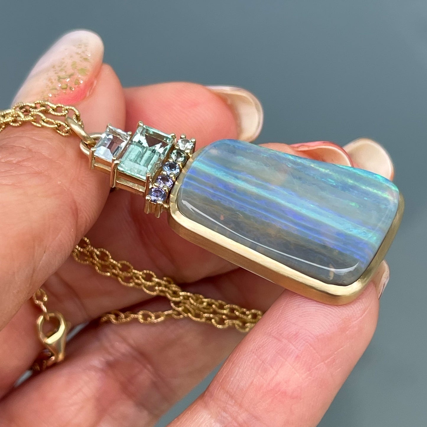 An Australian Opal Necklace by NIXIN Jewelry held sideways in front of a grey backdrop. Image displays the matte finish of the gold setting.