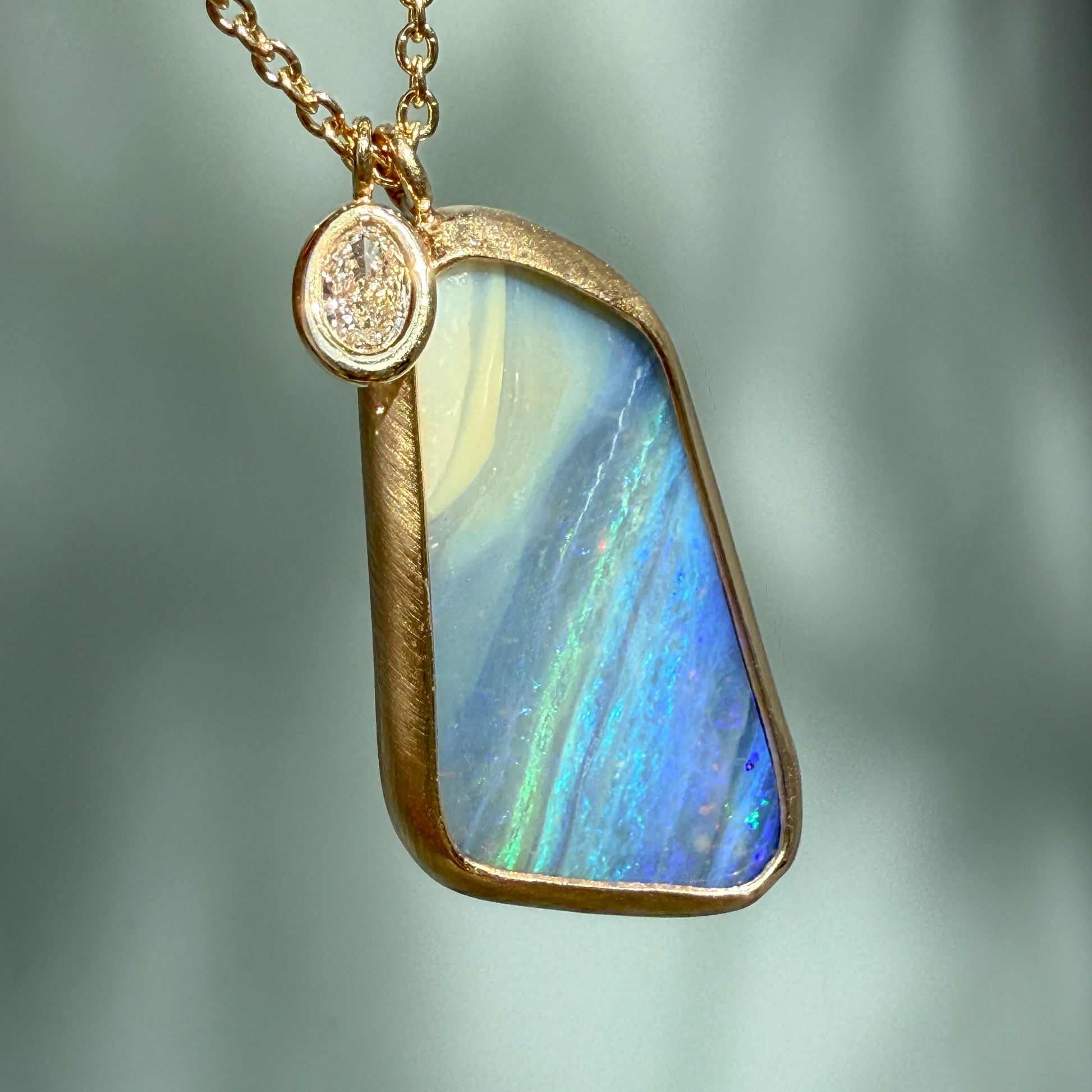 An Australian Opal Necklace by NIXIN Jewelry. A detail shot highlighting the textured, matte finish of the bezel setting next to the smooth, polished oval diamond. The unique opal necklace design emphasizes the contrast between the metal finishes and the oceanic opal stone.