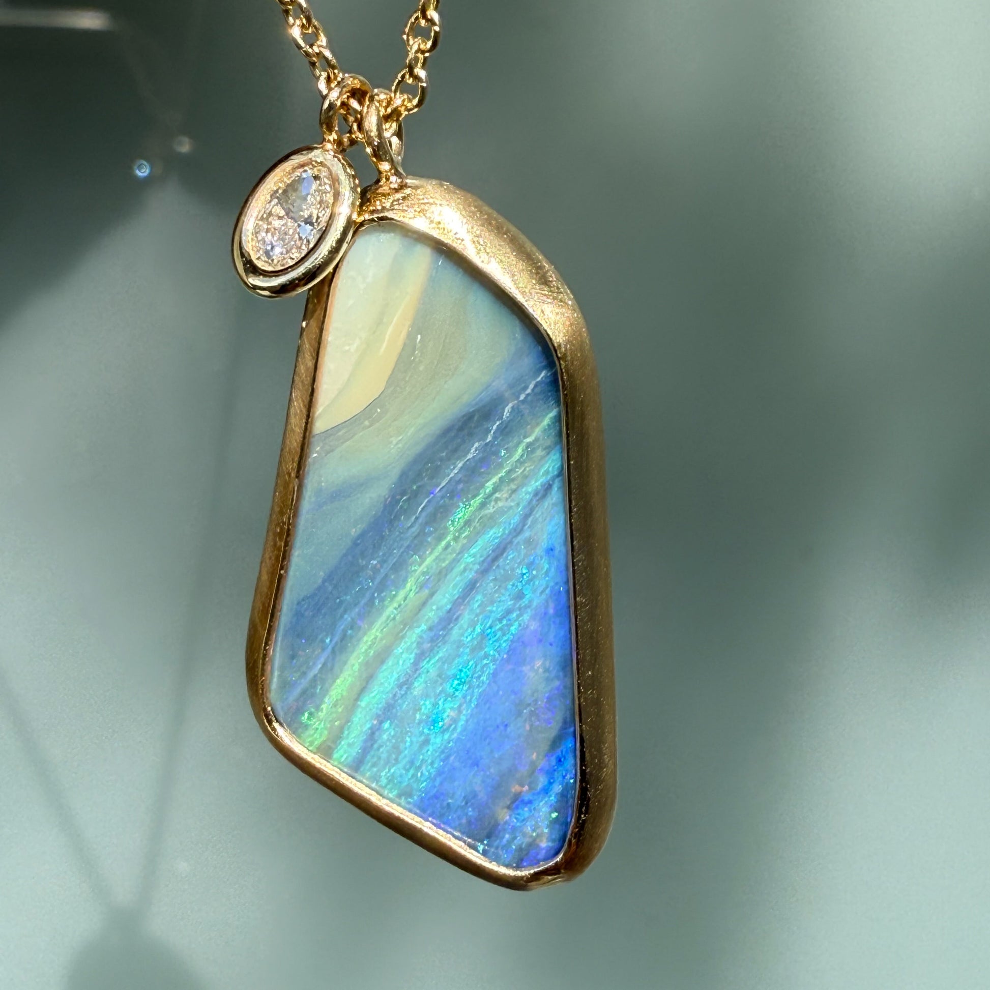 An Australian Opal Necklace by NIXIN Jewelry showcasing the striking blue color bar of the boulder opal against a matte gold bezel. This luxury necklace features a contrasting high-polish bezel set diamond charm. The piece reflects unique fine jewelry craftsmanship.