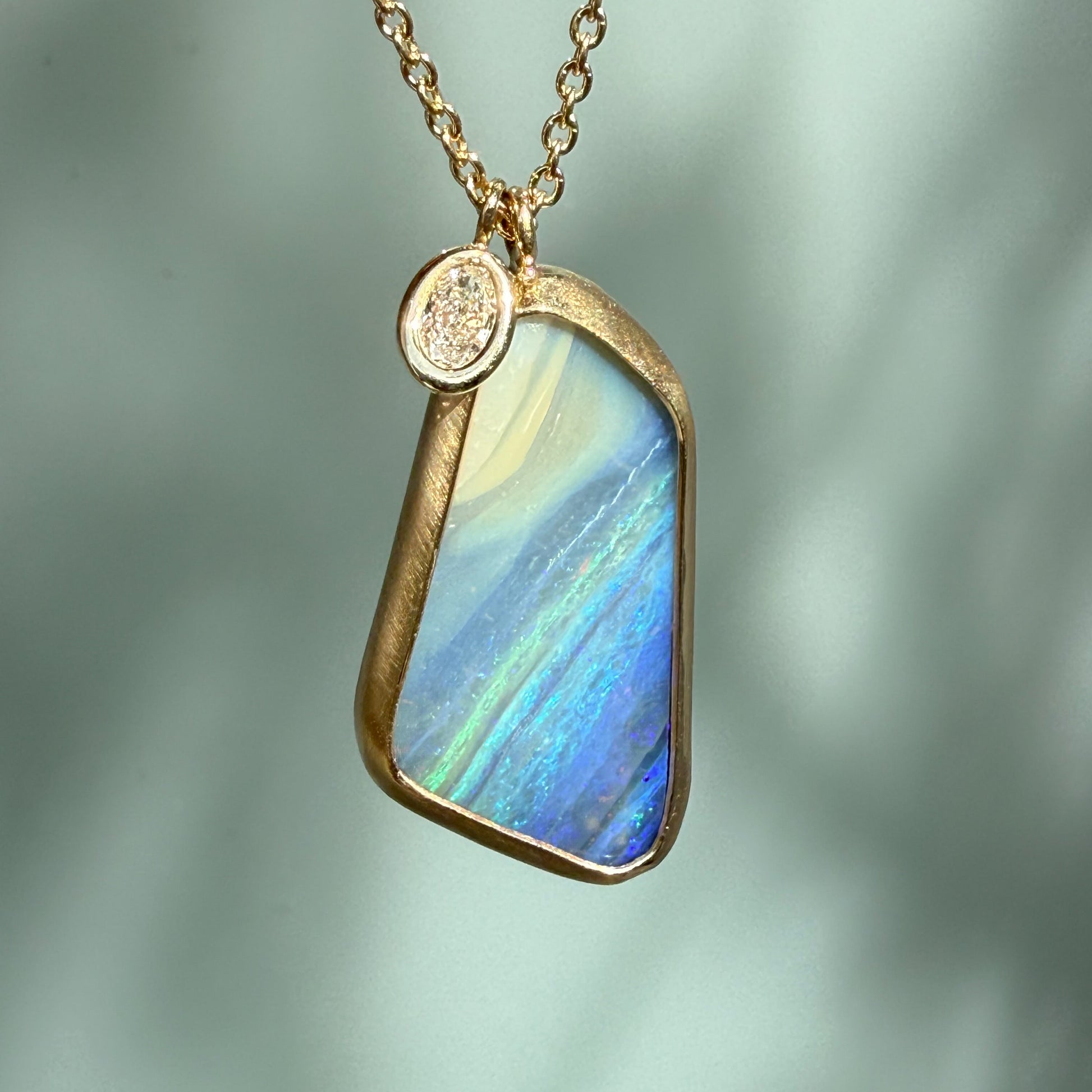 An Australian Opal Necklace by NIXIN Jewelry close-up showing the rich, striped blue and green hues of the natural opal in the gold opal necklace. The opal pendant design is jewelry art, pairing a rare opal jewelry stone with an oval diamond charm.