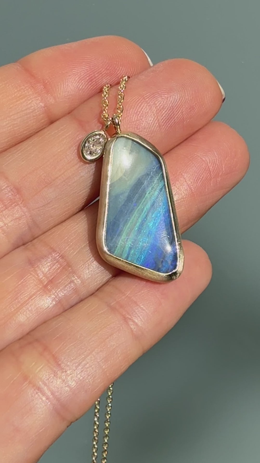 Video of an Australian Opal Necklace by NIXIN Jewelry showing the boulder opal pendant being gently turned to reveal the blue opal color shift and the textured ironstone back. This luxury jewelry video highlights the contrast between the polished diamond charm and the matte bezel setting, as well as the polished gold waves lapping the back of the opal stone.