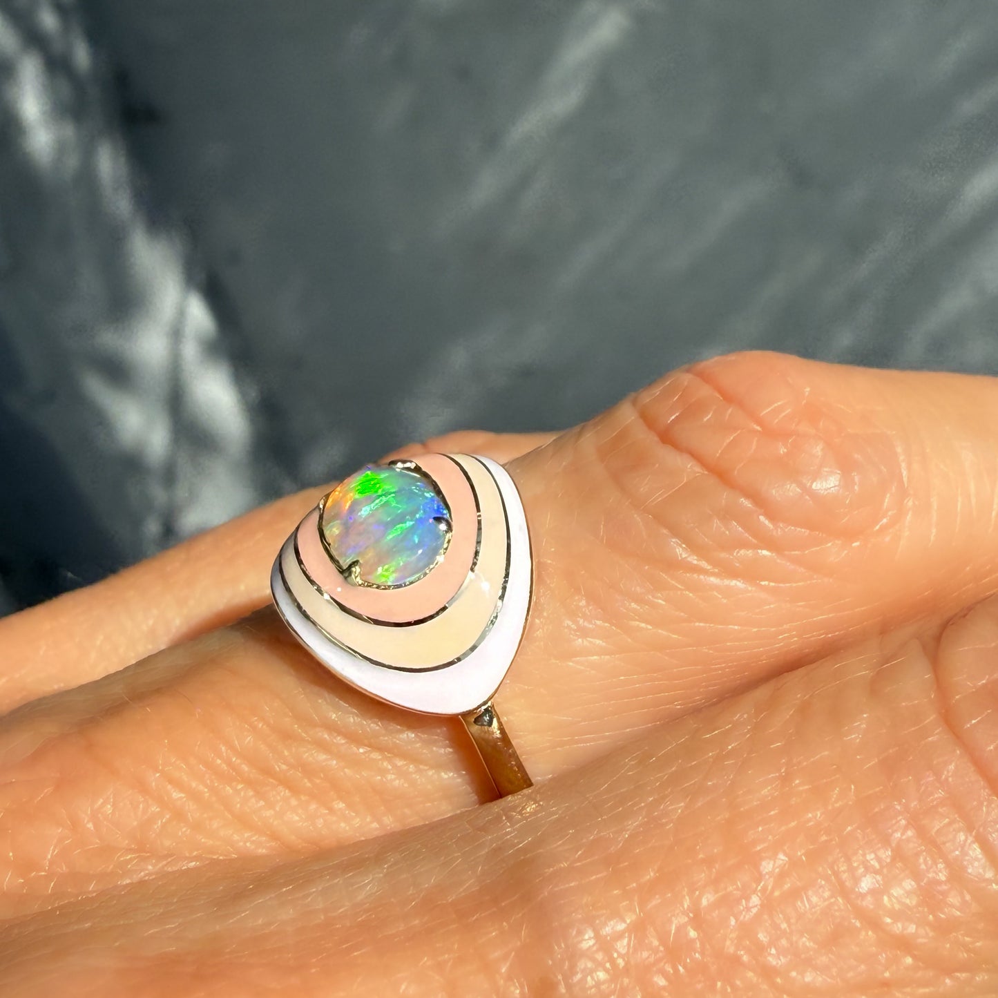 An Australian Opal Ring by NIXIN Jewelry, shown in profile, on a finger, against a gray backdrop. The central crystal opal emits vibrant flashes of color, set in a gold opal ring with a bold ombre enamel design.