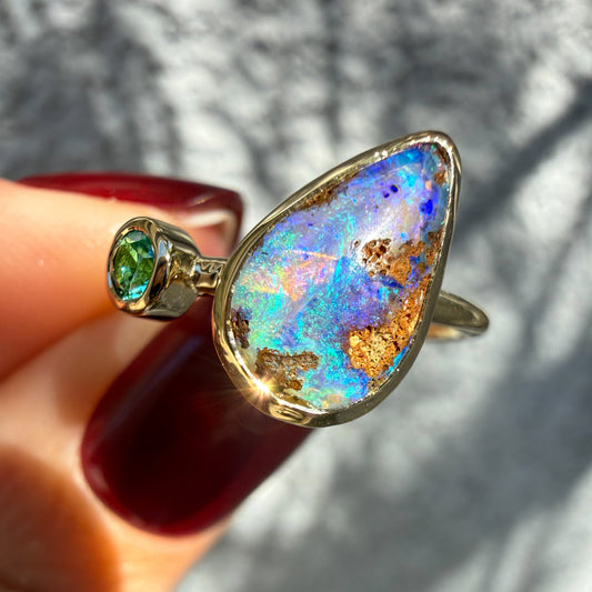 This close-up photograph provides a tilted perspective of an Australian Opal Ring by NIXIN Jewelry held between fingers with deep red nails. The view emphasizes the complex ironstone matrix embedded within the natural opal stone next to the vibrant green emerald. This dynamic pairing creates a piece of unique opal jewelry suitable as a cocktail ring.