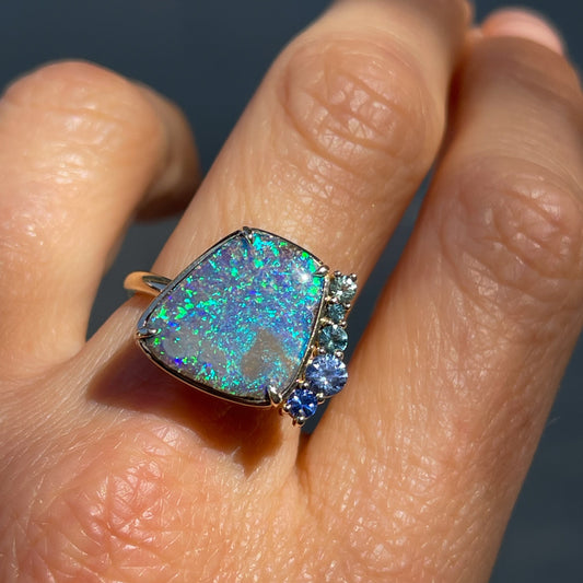 An Australian Opal Ring by NIXIN Jewelry modeled on a hand. Shows the blue boulder opal and matching teal sapphires.