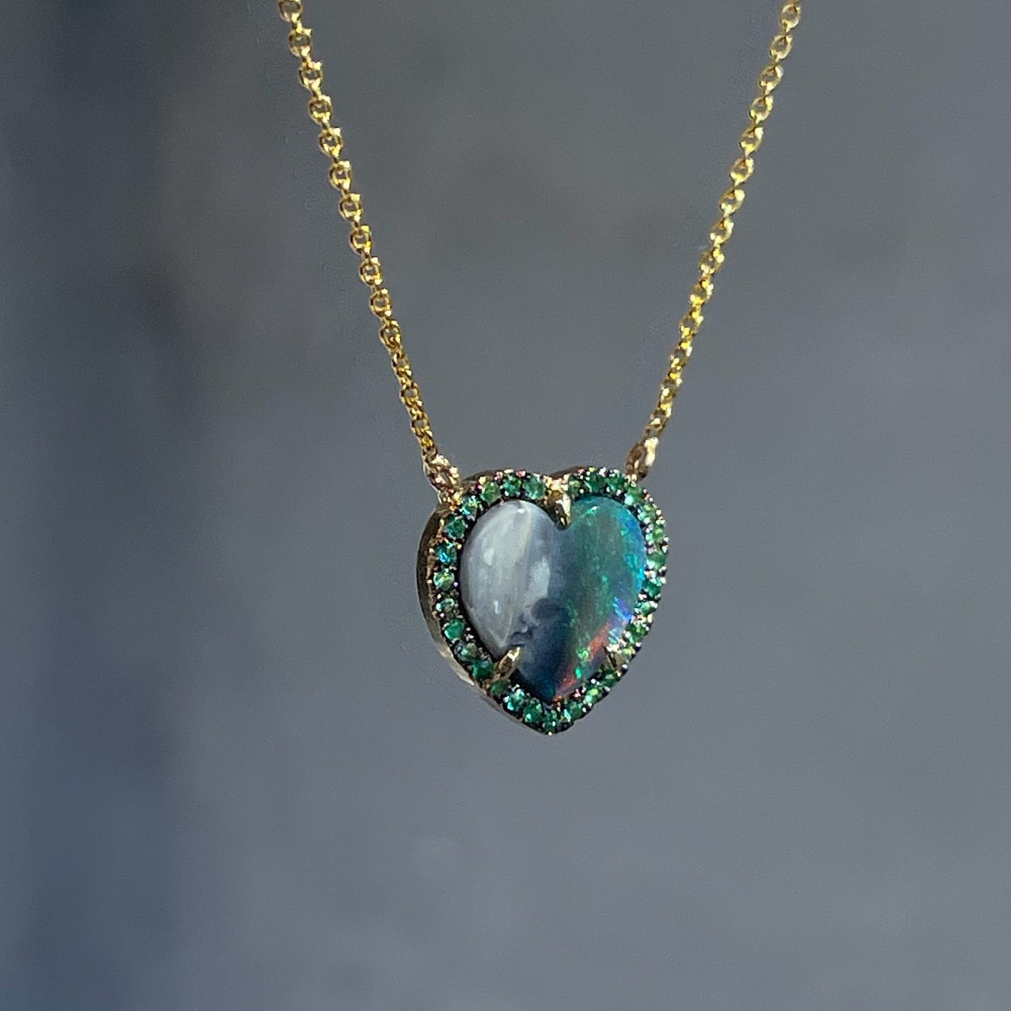 An Australian Opal Heart Necklace by NIXIN Jewelry hangs at a slight angle in the shade. A gold opal necklace made with emeralds and a Black Opal.
