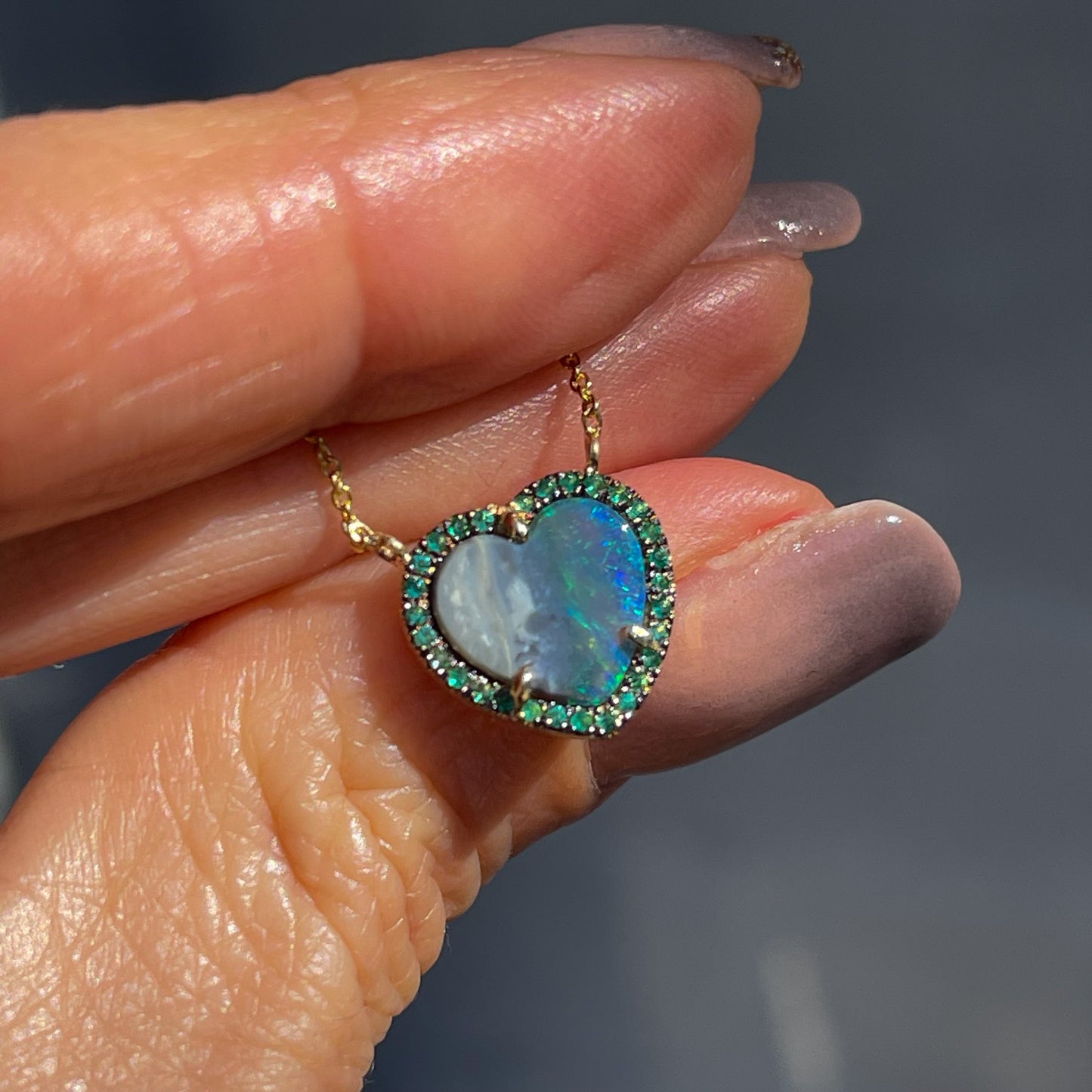 An Australian Opal Heart Necklace by NIXIN Jewelry poses on top of a hand. The opal heart is surrounded by emeralds.