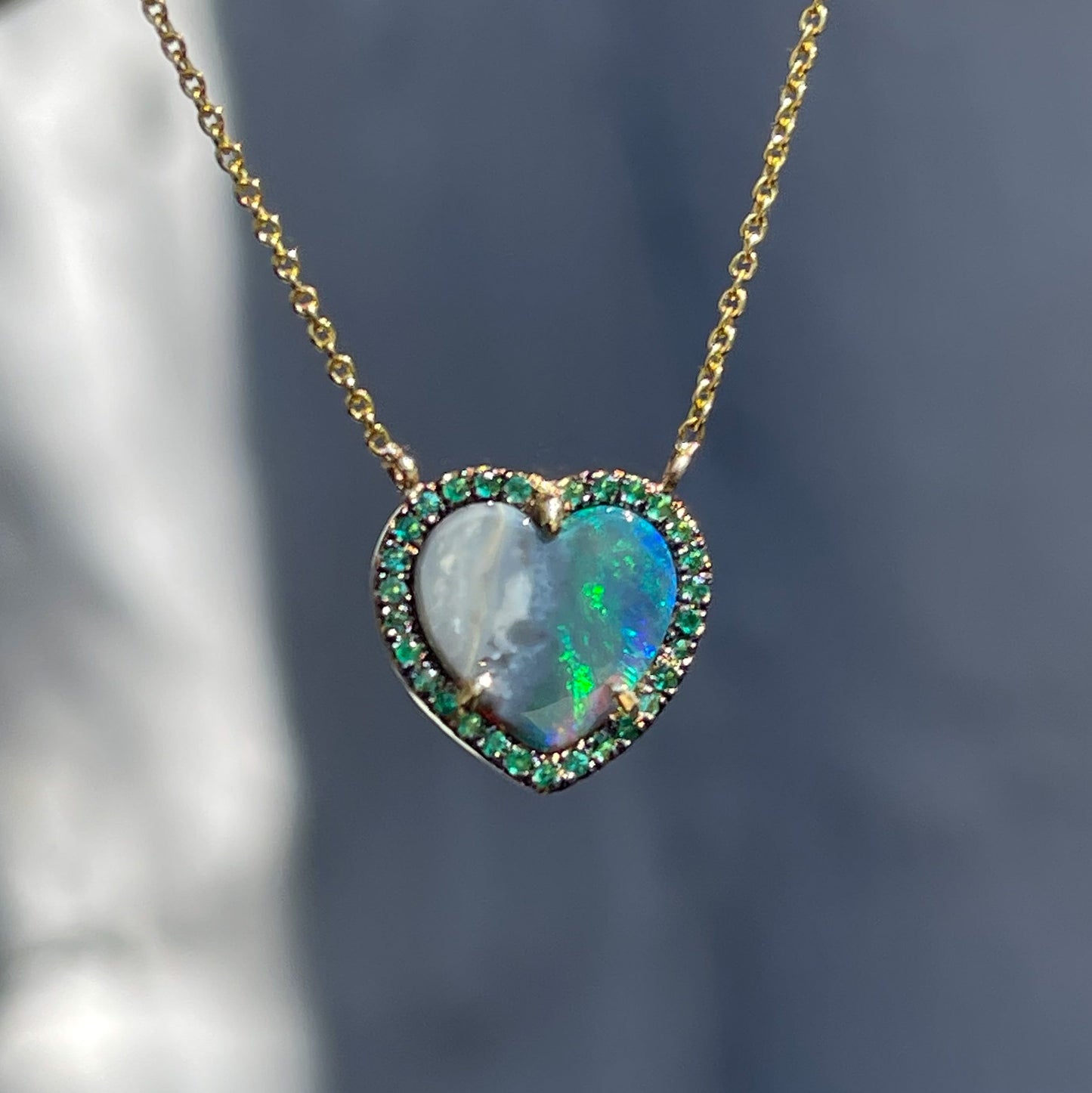 An Australian Opal Heart Necklace by NIXIN Jewelry hangs in sunlight. The blue green opal from Lightning Ridge is framed by emeralds and gold.