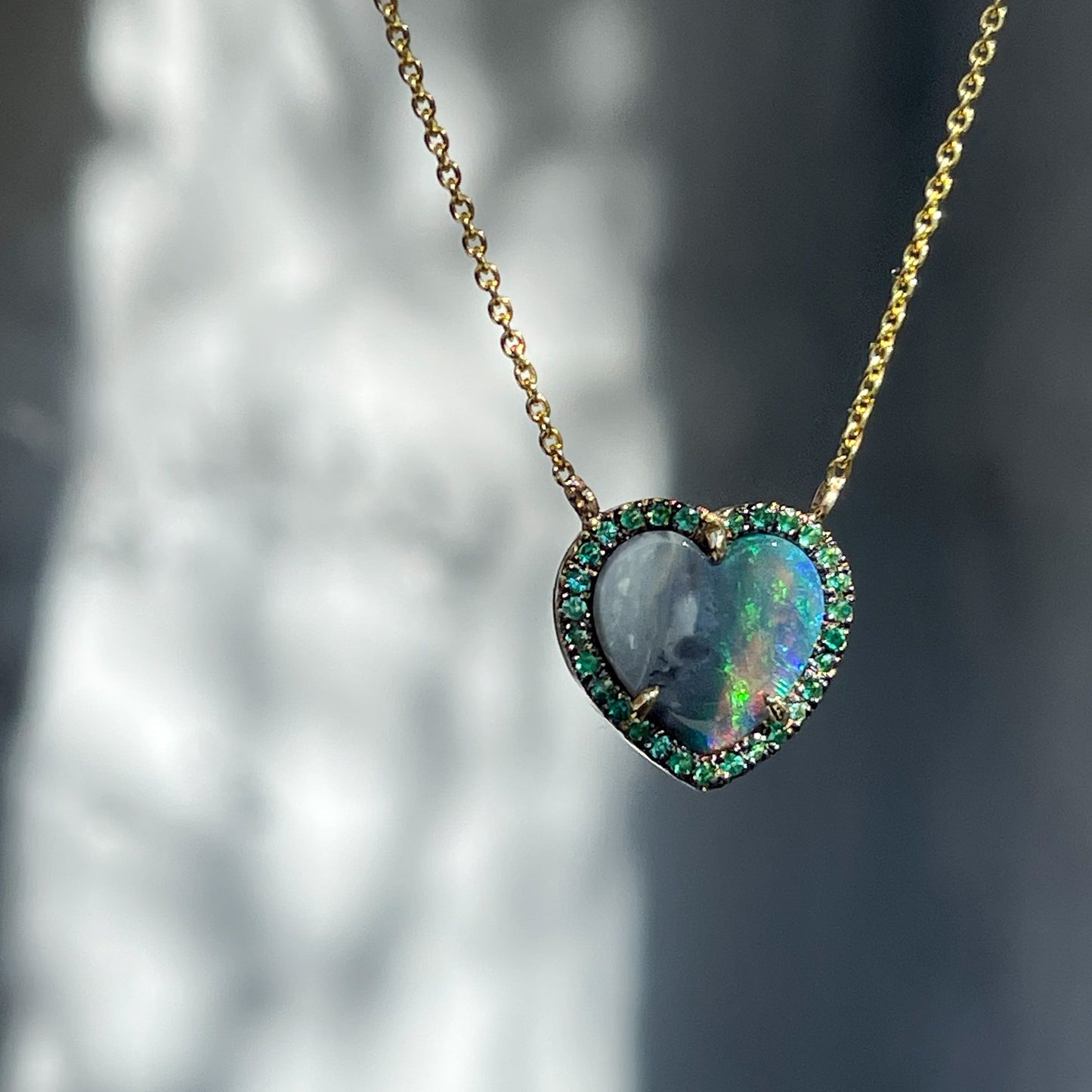 An Australian Opal Heart Necklace by NIXIN Jewelry hangs in front of a grey wall. An emerald an opal necklace in 14k gold.