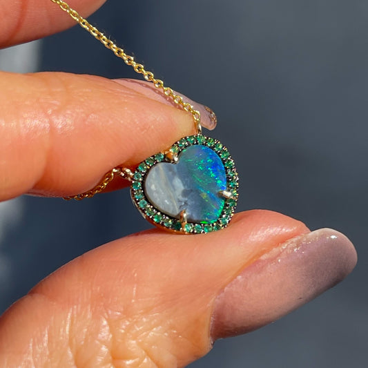 An Australian Opal Heart Necklace by NIXIN Jewelry held in sunlight. Made in 14k gold with a Black Opal and emeralds.