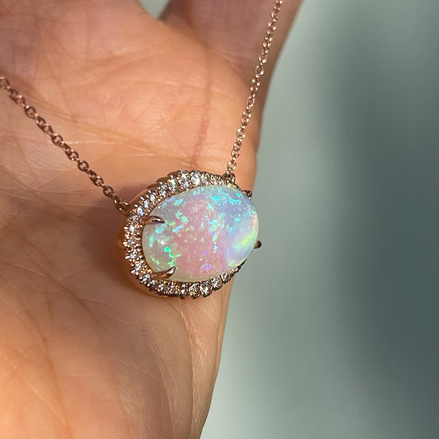 An Australian Opal Necklace by NIXIN Jewelry shot from the side, resting on a hand. The image displays the diamond halo and prong setting.