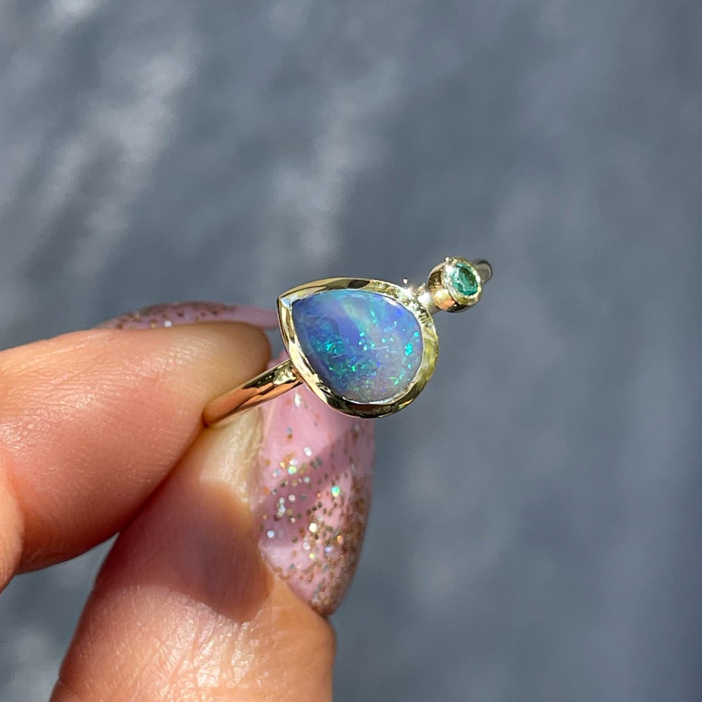 An Australian Opal Ring by NIXIN Jewelry held in front of a gray backdrop. A Lightning Ridge Black Opal Ring with a bezel set emerald.