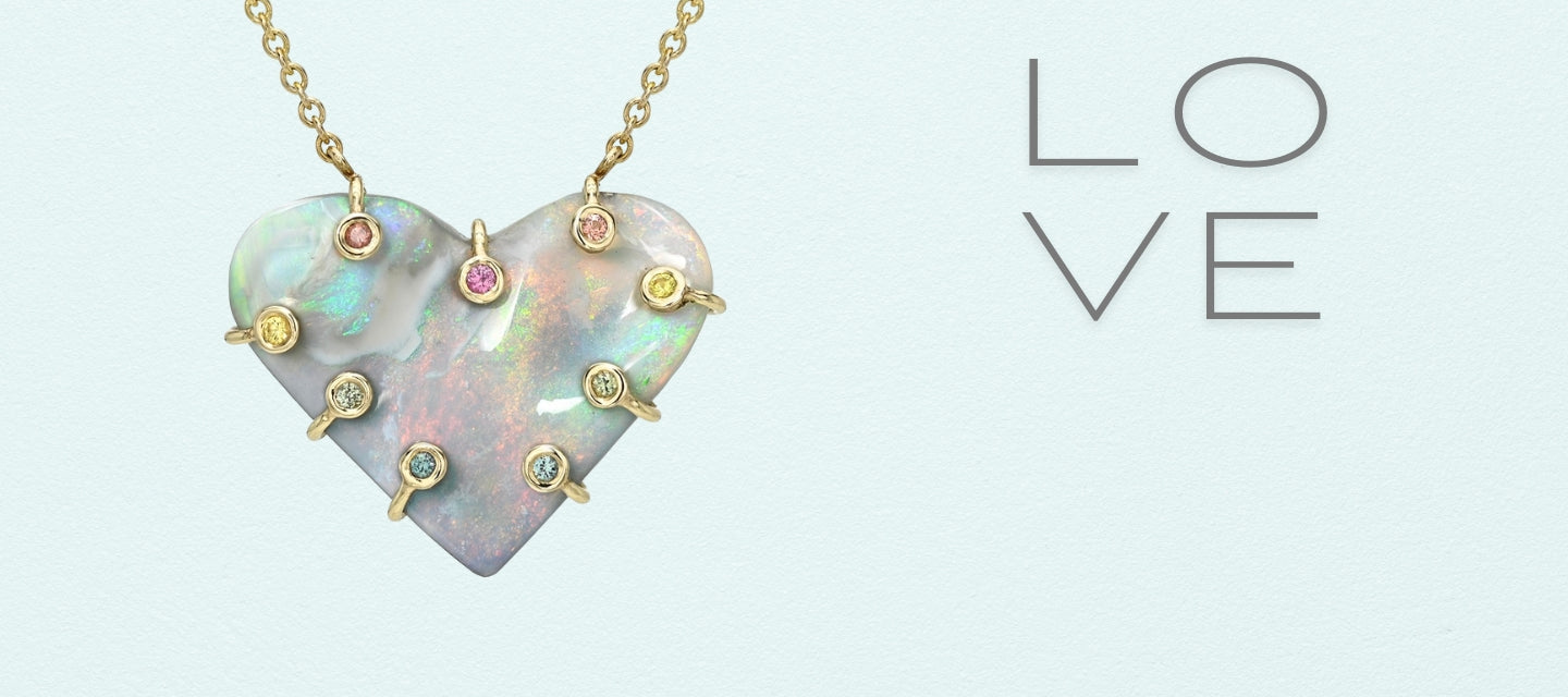 An Australian Opal Necklace by NIXIN Jewelry hanging in front of a blue backdrop. An opal heart necklace with sapphire accents set in 14k gold.