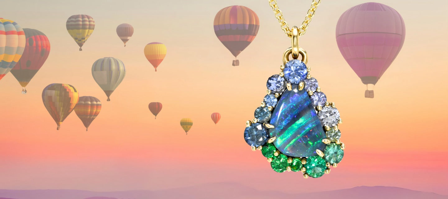 An Australian Opal Necklace by NIXIN Jewelry staged in front of a backdrop of ascending hot air balloons. The opal pendant is surrounded by a sapphire halo, punctuate with emeralds and garnets.