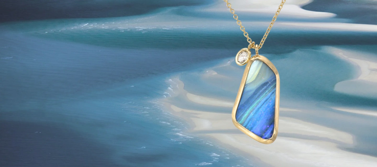 An Australian Opal Necklace by NIXIN Jewelry with a Boulder Opal in a gold bezel setting. The gold opal necklace has a diamond charm and hangs in front of a blue water background