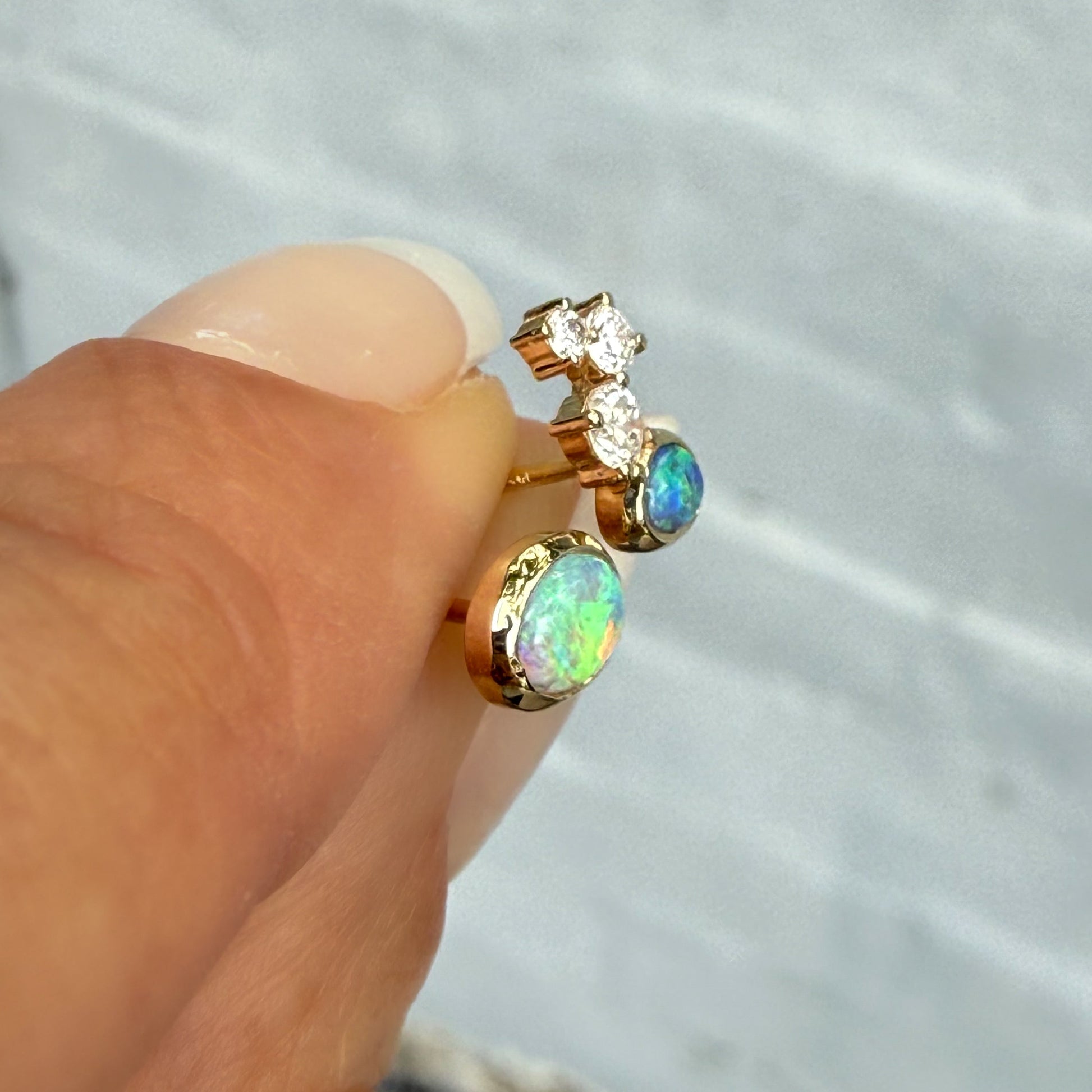 Australian Opal Earrings by NIXIN Jewelry. Handheld side view of the 14k gold opal earrings, emphasizing the vivid green and blue flash of the black opal jewelry. These mismatched earrings are unique opal earrings perfect for luxury jewelry buyers.