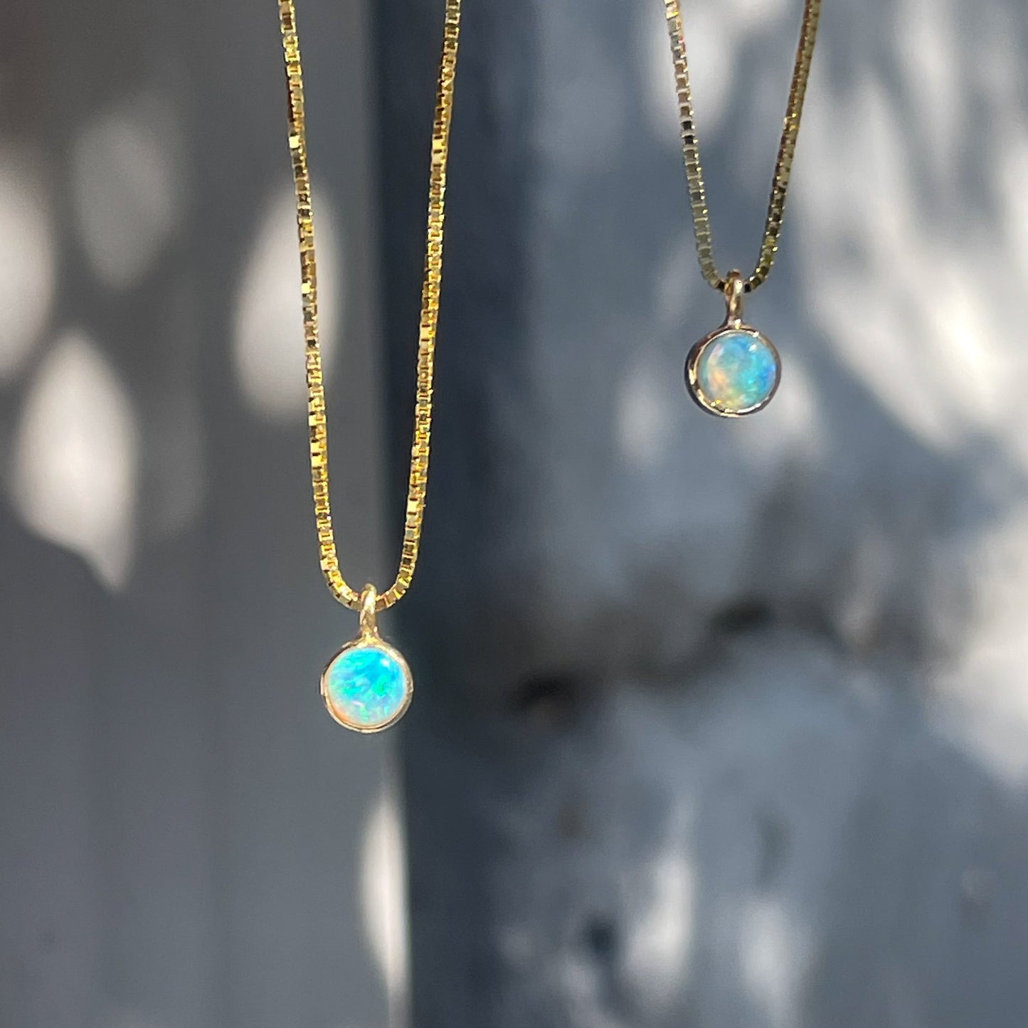 An Australian Opal Necklaces design by NIXIN Jewelry. Two of the opal necklaces hang in front of a grey wall with small blue opals in the pendants.