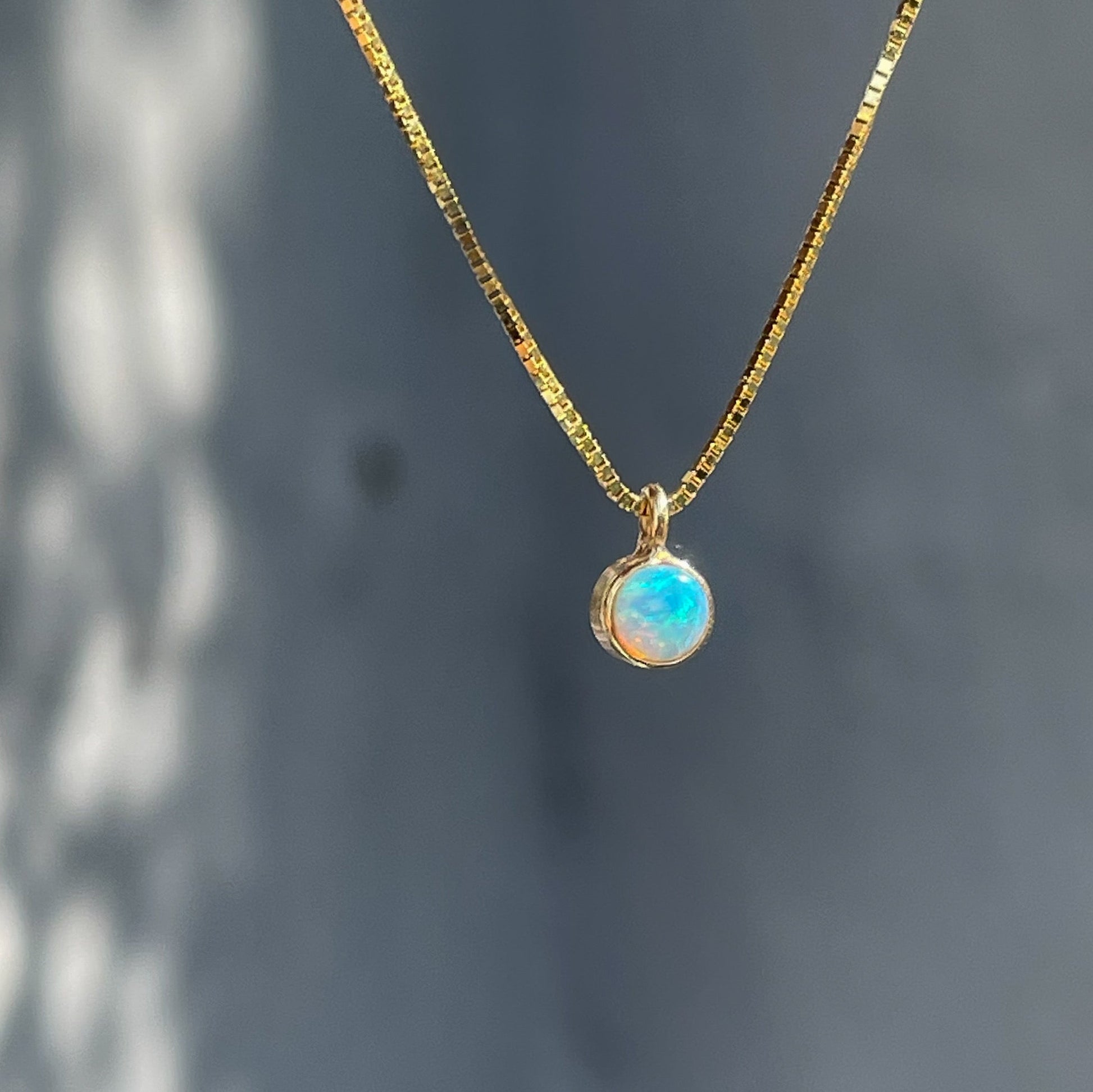 An Australian Opal Necklace by NIXIN Jewelry hangs in front of a grey wall. The dainty opal necklace is made in 14k gold.