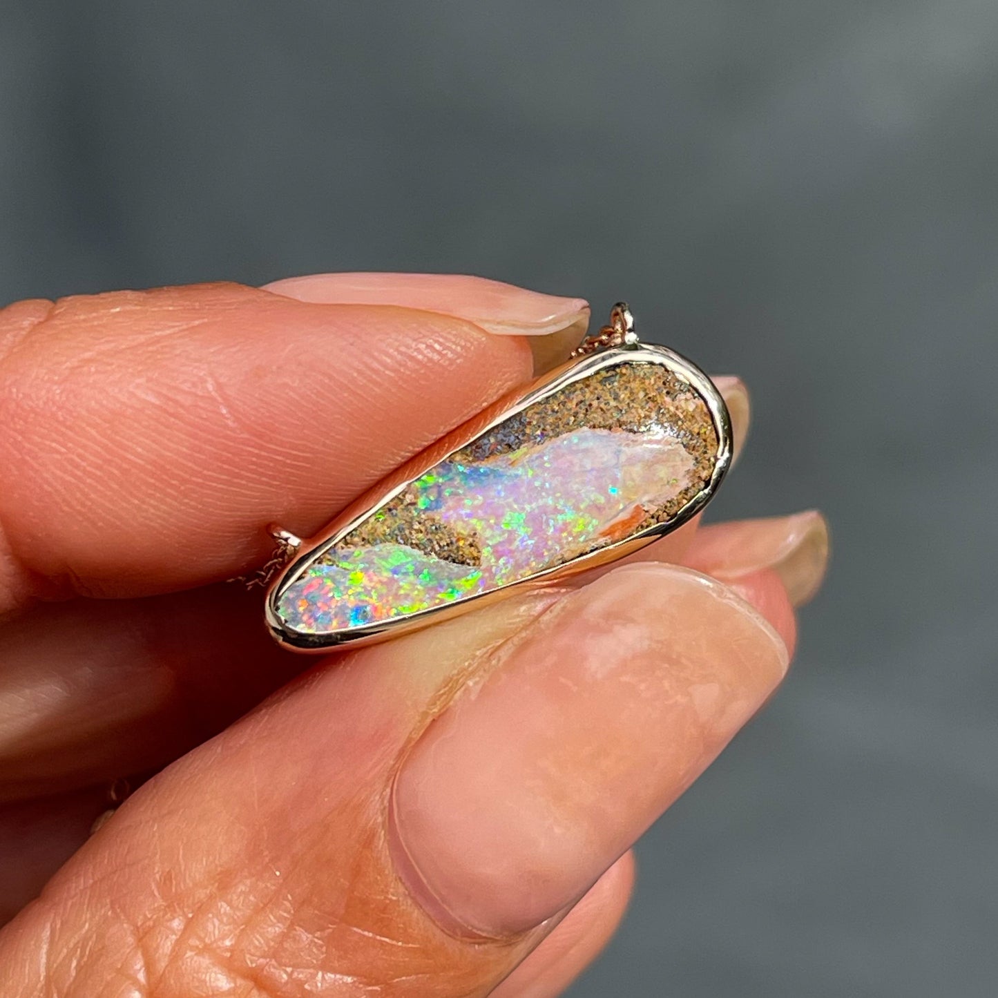 An Australian Opal Necklace by NIXIN Jewelry held in the sunlight. The opal stone flashes pink, purple, green and blue and is set in 14k rose gold.