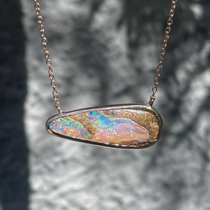 An Australian Opal Necklace by NIXIN Jewelry hangs in front of a grey wall. The natural opal flashes pink and blue and other colors of the rainbow.