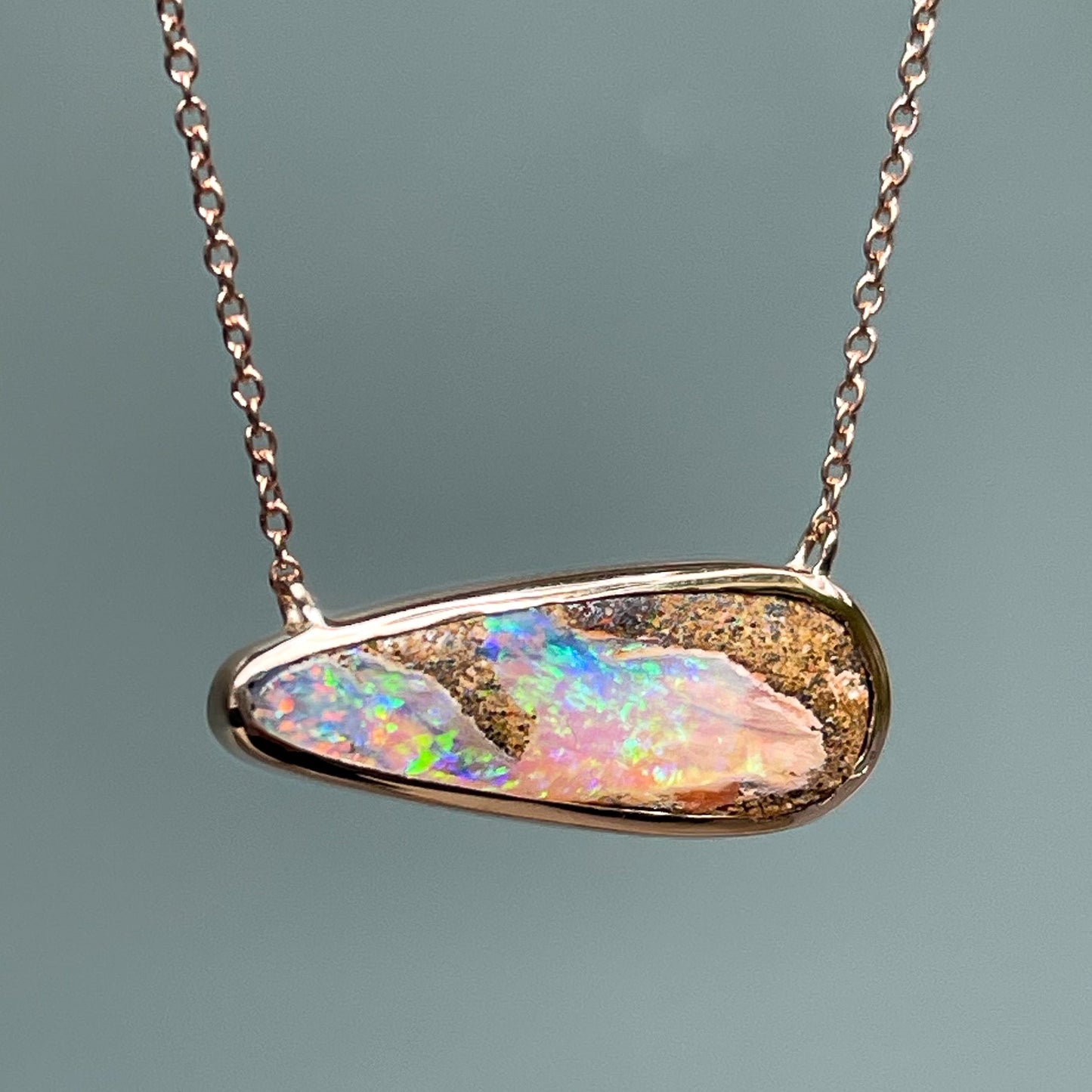 An Australian Opal Necklace by NIXIN Jewelry hangs in front of a frosted glass backdrop. The pipe opal is set in a rose gold bezel setting.