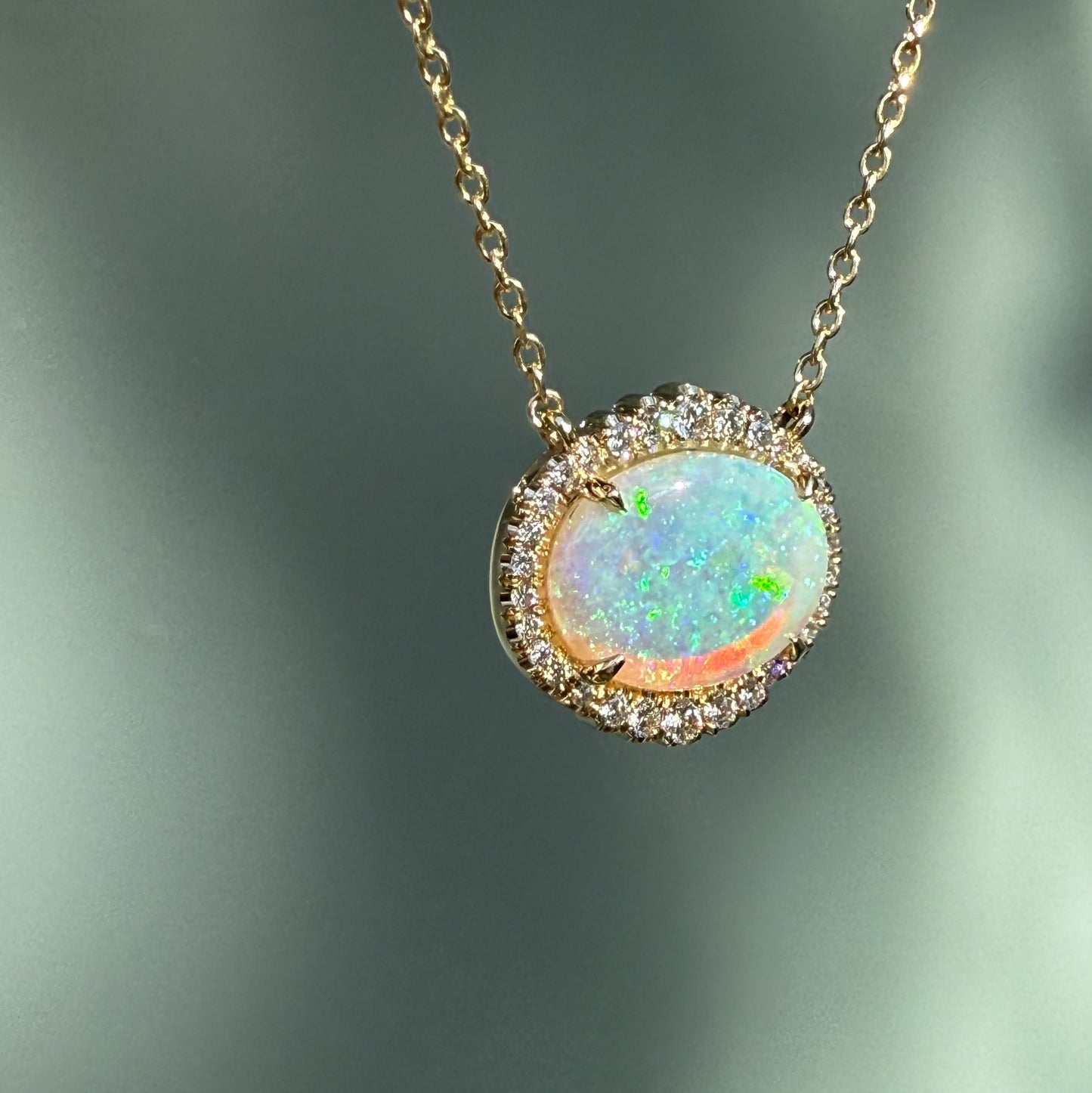 An Australian Opal Necklace by NIXIN Jewelry, highlighting the intense play-of-color in a natural opal. This unique opal jewelry piece features an oval opal stone with a shimmering diamond halo, hanging at an angle. An exquisite women's luxury necklace and rare opal jewelry offering.