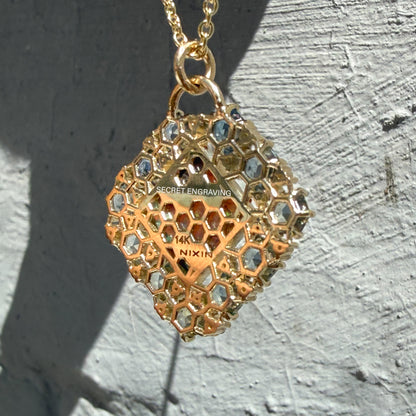 The intricate reverse side reveals the hidden details of an Australian Opal Pendant by NIXIN Jewelry against a grey background. This view of the unique opal necklace highlights an open honeycomb latticework in solid 14k gold, complete with text pointing out the location of a secret engraving. The geometric gold framework demonstrates the level of high art and design involved in this custom piece of unique opal jewelry.
