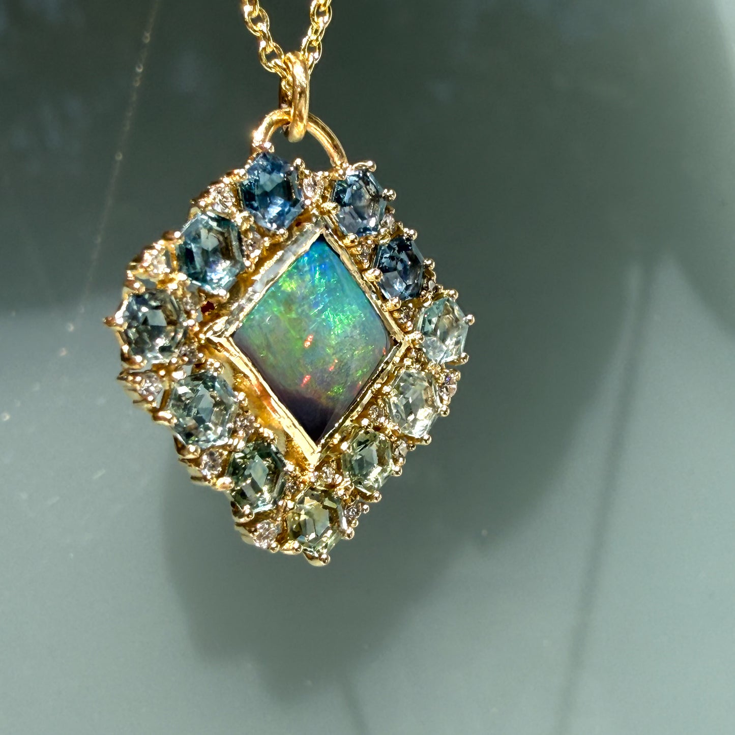 A dramatic angled shot illuminates an Australian Opal Pendant by NIXIN Jewelry hanging against a glass background. This specific view of the black opal necklace emphasizes the sudden red and orange fire flashing from the precious opal center. A solid bezel secures the stone, bordered by a custom layout of geometric sapphires that define this unique jewelry piece.