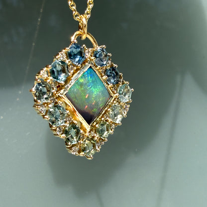 A dramatic angled shot illuminates an Australian Opal Pendant by NIXIN Jewelry hanging against a glass background. This specific view of the black opal necklace emphasizes the sudden red and orange fire flashing from the precious opal center. A solid bezel secures the stone, bordered by a custom layout of geometric sapphires that define this unique jewelry piece.