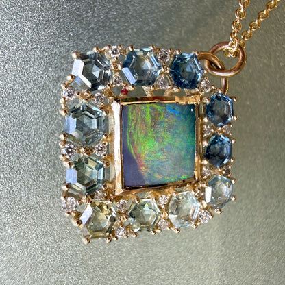 A close-up macro photograph features an Australian Opal Pendant by NIXIN Jewelry. This rare opal jewelry design showcases a square lightning ridge opal with a high polish finish inside a protective bezel setting. The central black opal is encircled by a geometric ombre jewelry halo of hexagon-cut montana sapphires and tiny diamonds.