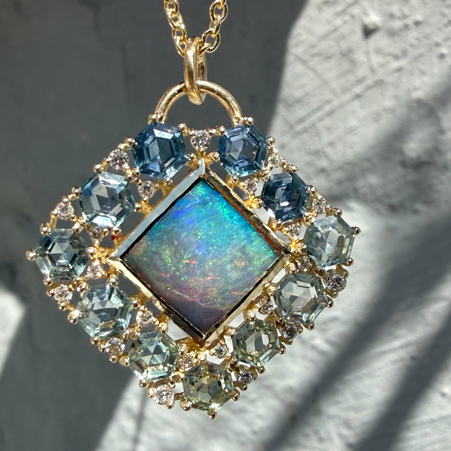 A macro view highlights an Australian Opal Pendant by NIXIN Jewelry. This piece of unique fine jewelry showcases a natural opal center stone displaying vivid neon blue and green hues. The central black opal is framed by a geometric ombré jewelry border composed of hexagon sapphire jewelry prong-set in yellow gold.