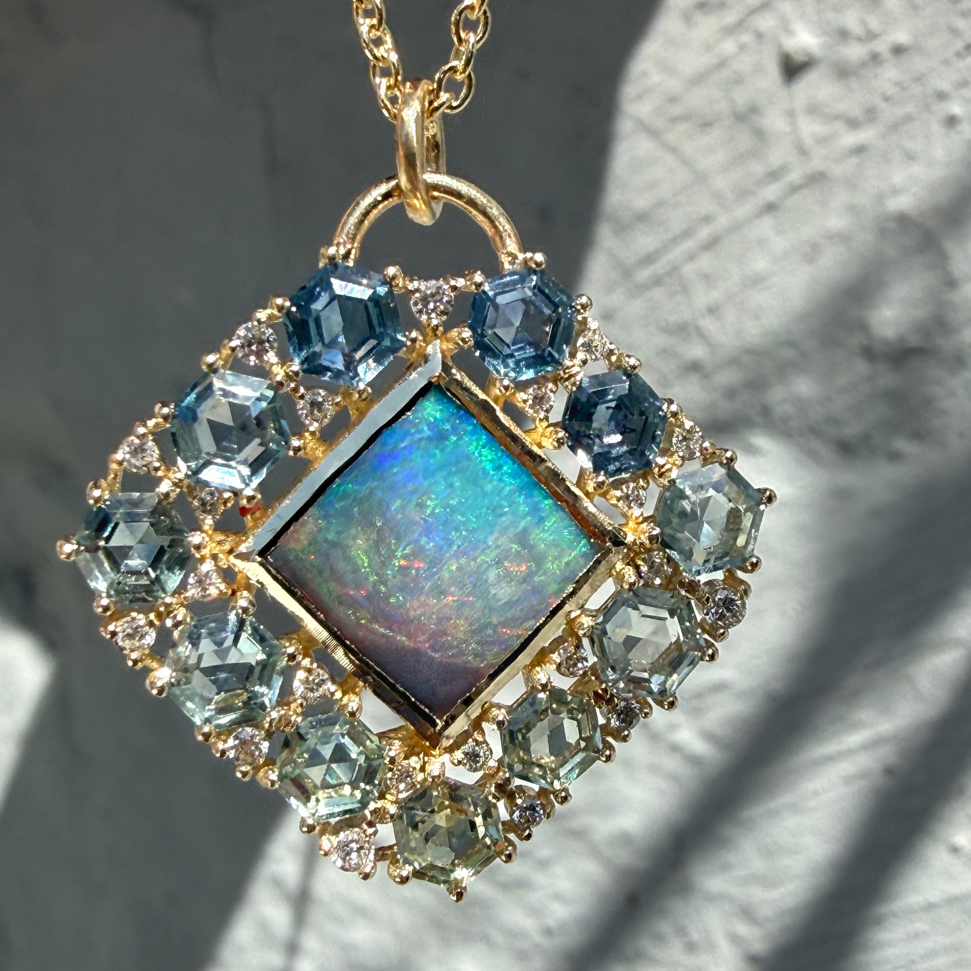 A macro view highlights an Australian Opal Pendant by NIXIN Jewelry. This piece of unique fine jewelry showcases a natural opal center stone displaying vivid neon blue and green hues. The central black opal is framed by a geometric ombré jewelry border composed of hexagon sapphire jewelry prong-set in yellow gold.