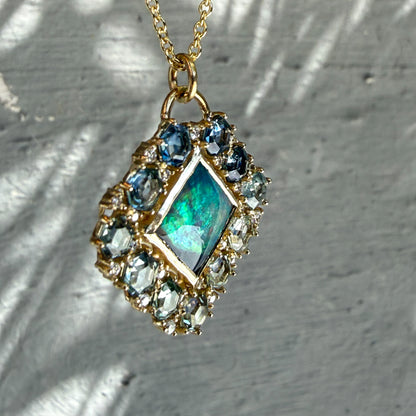 Sunbeams illuminate an Australian Opal Pendant by NIXIN Jewelry. This rare opal necklace features a distinct green and blue black opal jewelry center stone that shifts in changing light. The unique opal jewelry design is completed with a custom frame of hexagon-cut montana sapphires transitioning through deep teal and seafoam hues.