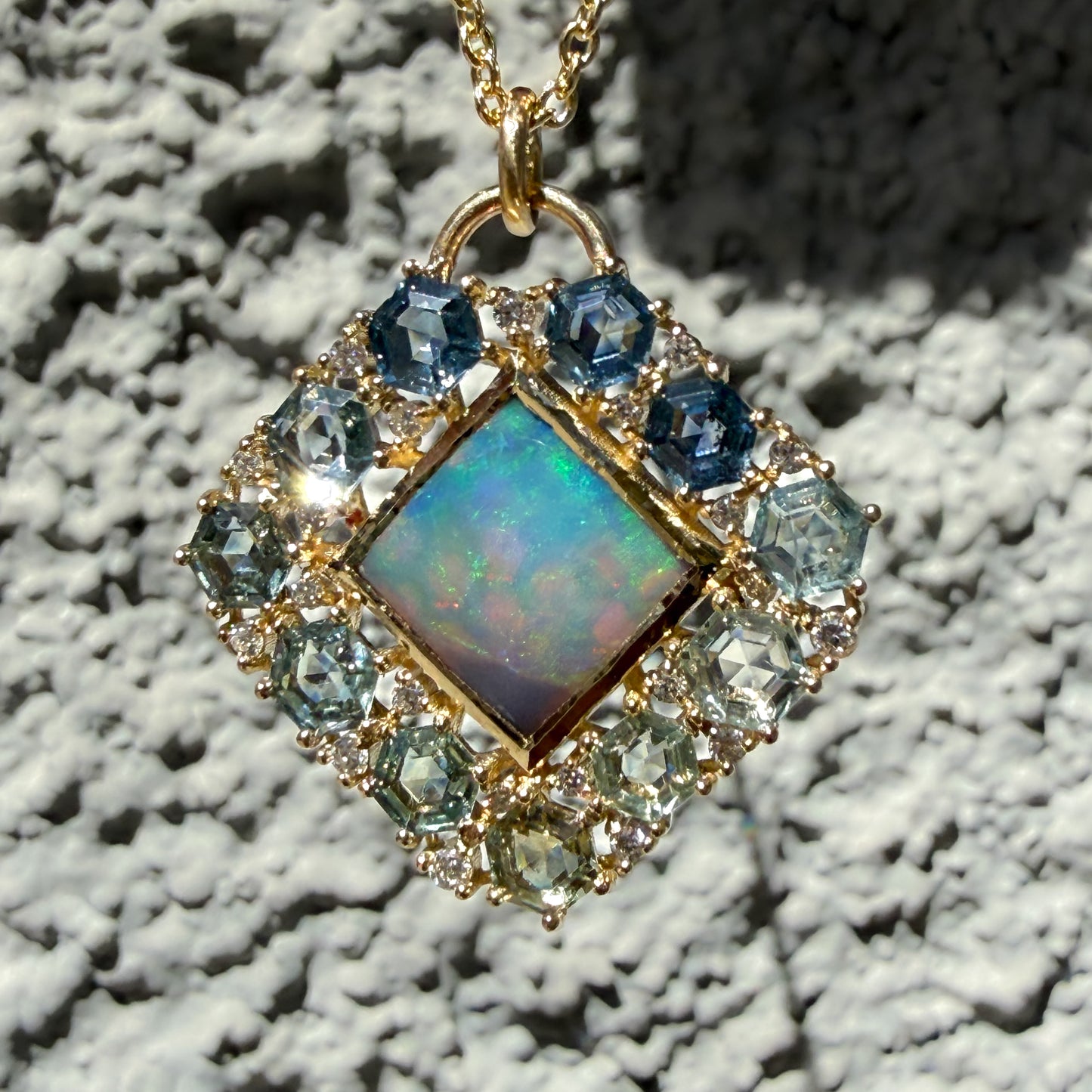 Hanging against a textured stone wall is an Australian Opal Pendant by NIXIN Jewelry. This luxury jewelry piece acts as a statement black opal necklace with its signature diamond-shaped silhouette and polish finish. The center stone is bordered by a structured layout of gradient sapphires and small accent diamonds to maximize its unique jewelry appeal.