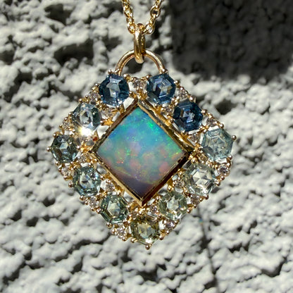 Hanging against a textured stone wall is an Australian Opal Pendant by NIXIN Jewelry. This luxury jewelry piece acts as a statement black opal necklace with its signature diamond-shaped silhouette and polish finish. The center stone is bordered by a structured layout of gradient sapphires and small accent diamonds to maximize its unique jewelry appeal.