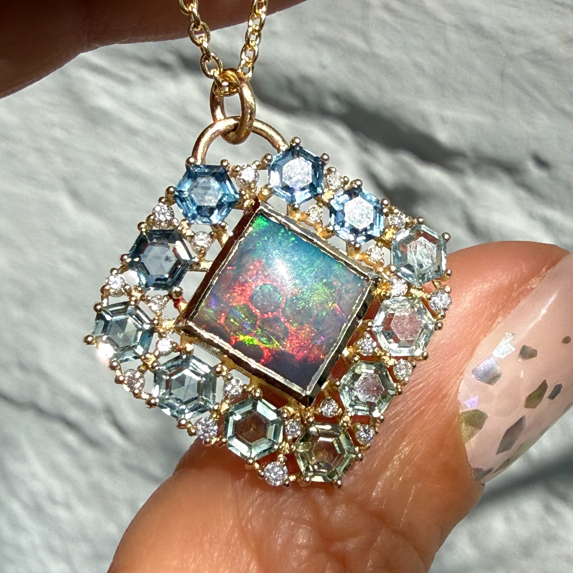 A hand displays an Australian Opal Pendant by NIXIN Jewelry. This women's luxury necklace features a rare lightning ridge opal that exhibits vibrant red, blue, and green play-of-color. The precious opal is secured in a polished 14k gold bezel setting and surrounded by a halo of hexagon-cut montana sapphires and prong-set diamonds.