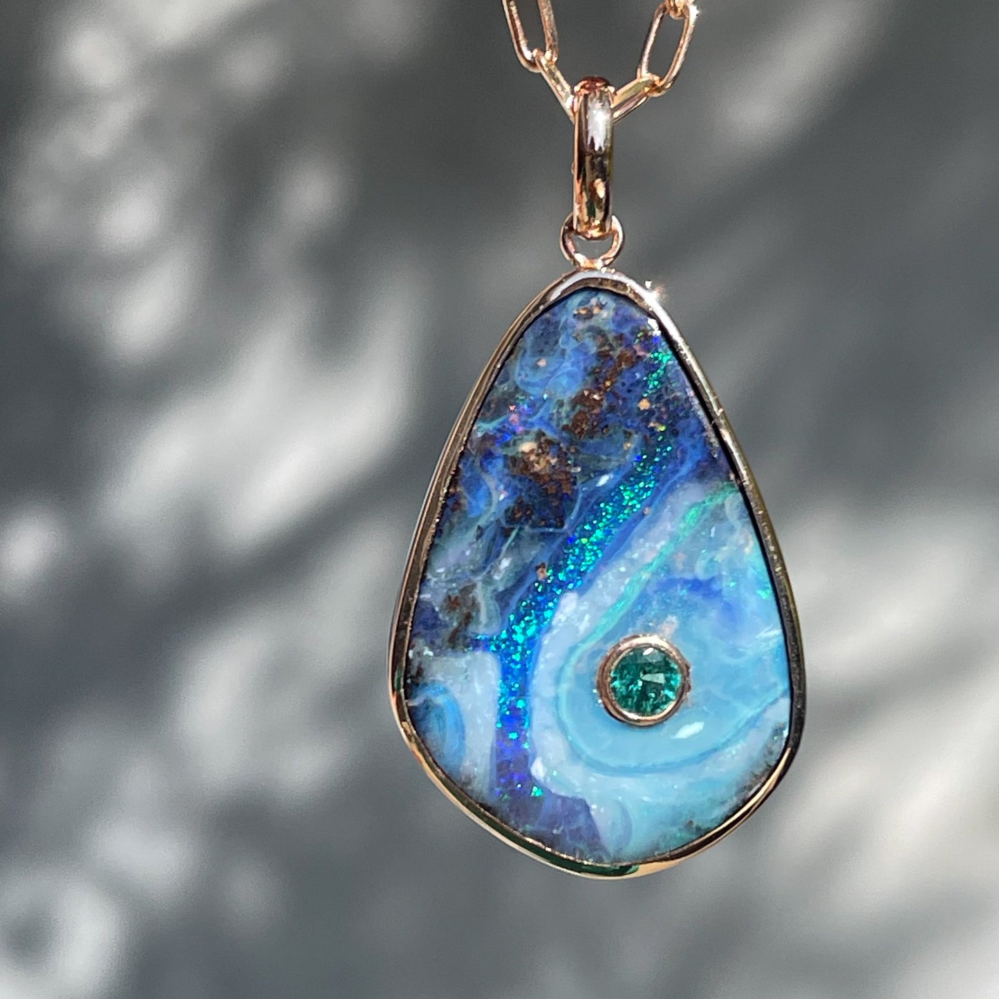 An Australian Opal Necklace by NIXIN Jewelry hanging in front of a grey backdrop. Made in rose gold with a bezel setting and hanging from a paperclip chain.