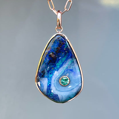 An Australian Opal Necklace by NIXIN Jewelry hanging in the shade in front of a frosted glass background. This piece of rare jewelry features a blue opal and an emerald set in polished gold.