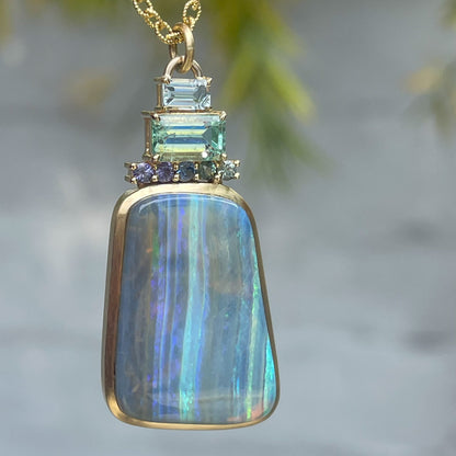 An Australian Opal Necklace by NIXIN Jewelry hangs in front of a grey wall. The blue opal blends well with the blue and green sapphires, aquamarine and emerald.