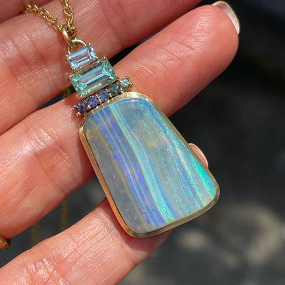 An Australian Opal Necklace by NIXIN Jewelry resting on a hand in daylight. The opal pendant is accented with sapphires and emerald.