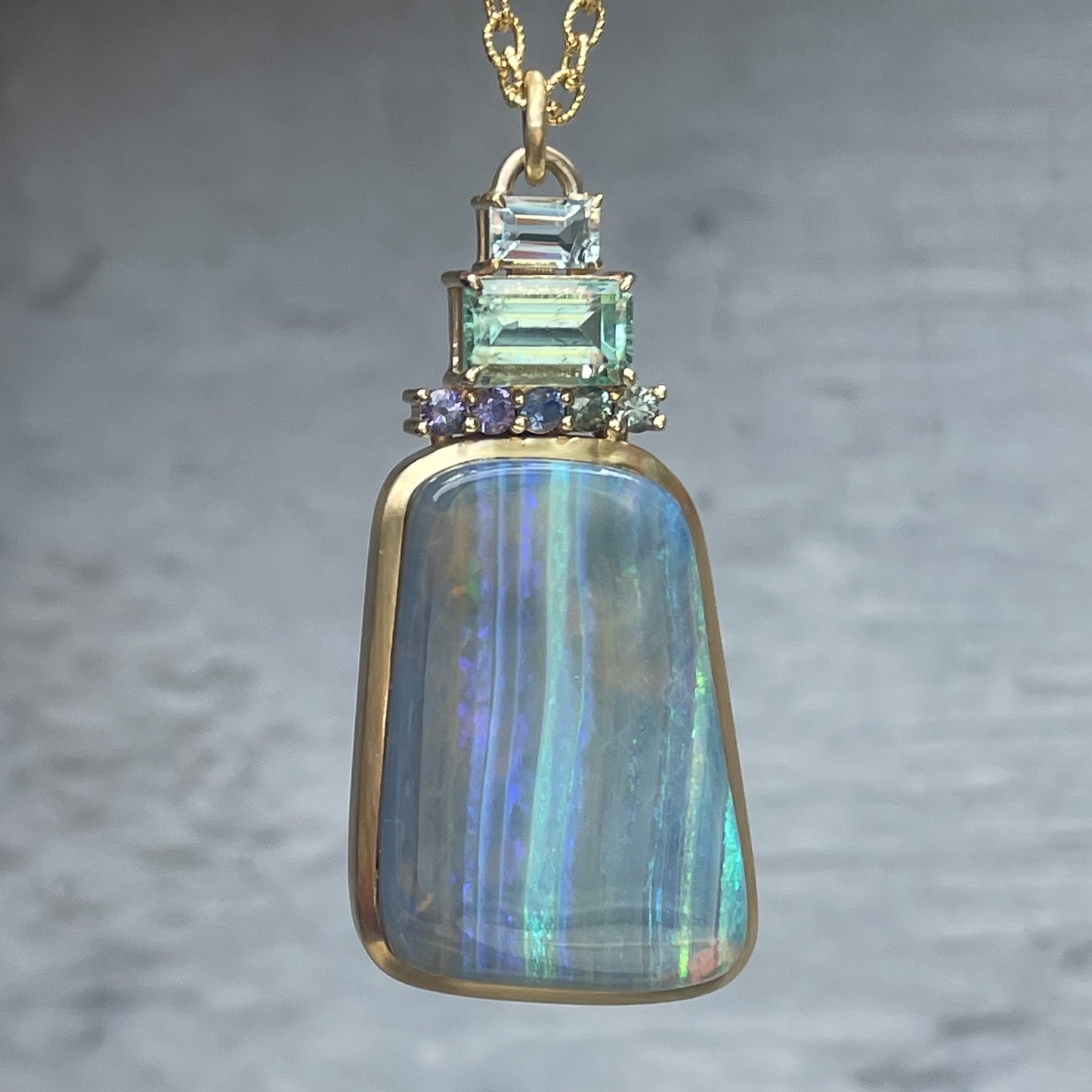 An Australian Opal Necklace by NIXIN Jewelry hangs in front of a grey wall. Atop the Blue Opal stack ombré sapphires, an emerald, and an aquamarine.
