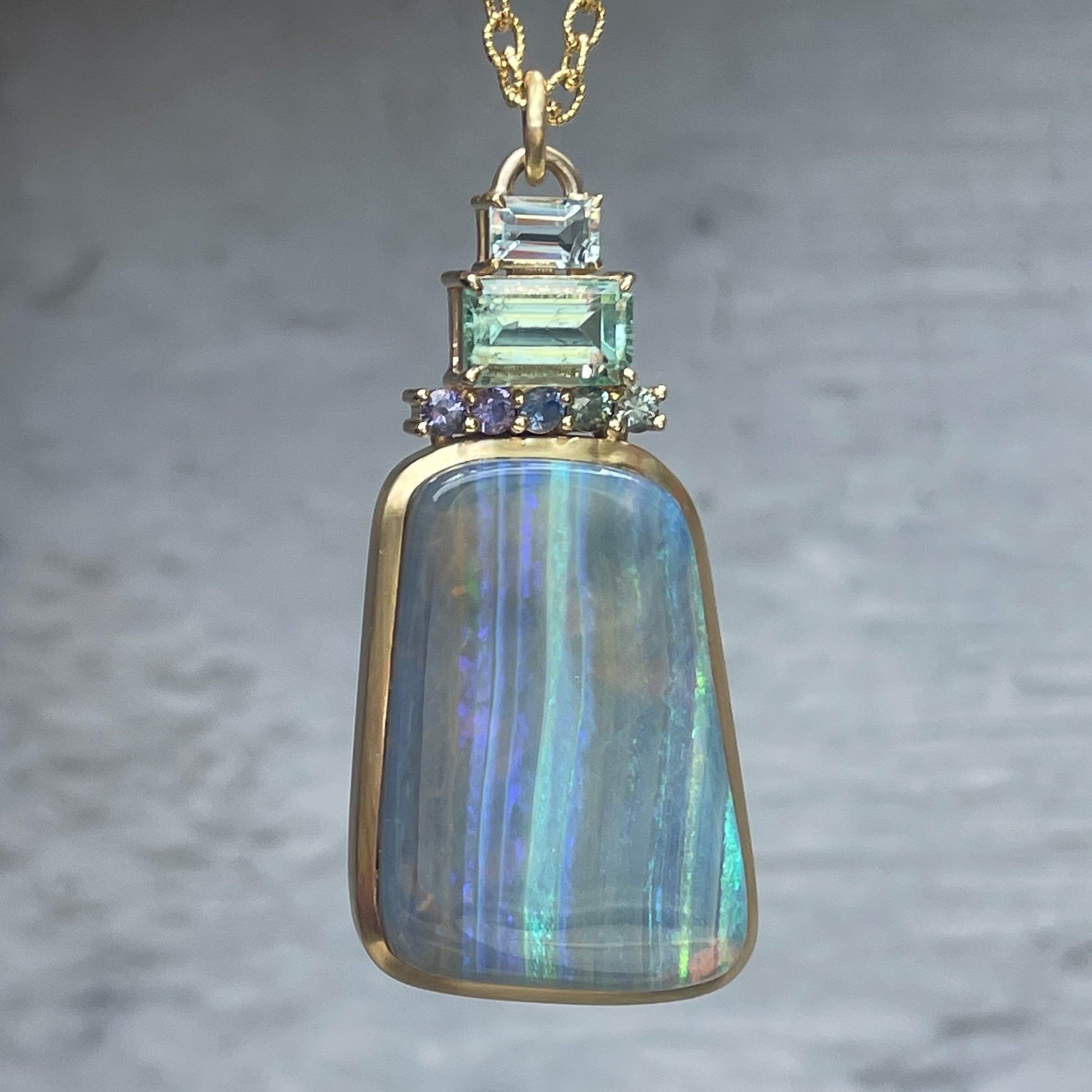 An Australian Opal Necklace by NIXIN Jewelry hangs in front of a grey wall. Atop the Blue Opal stack ombré sapphires, an emerald, and an aquamarine.