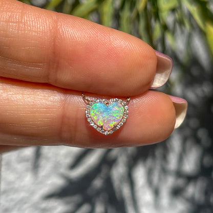 An Opal Heart Necklace by NIXIN Jewelry held between two fingers. The Australian opal is surrounded by a diamond halo and set in rose gold.