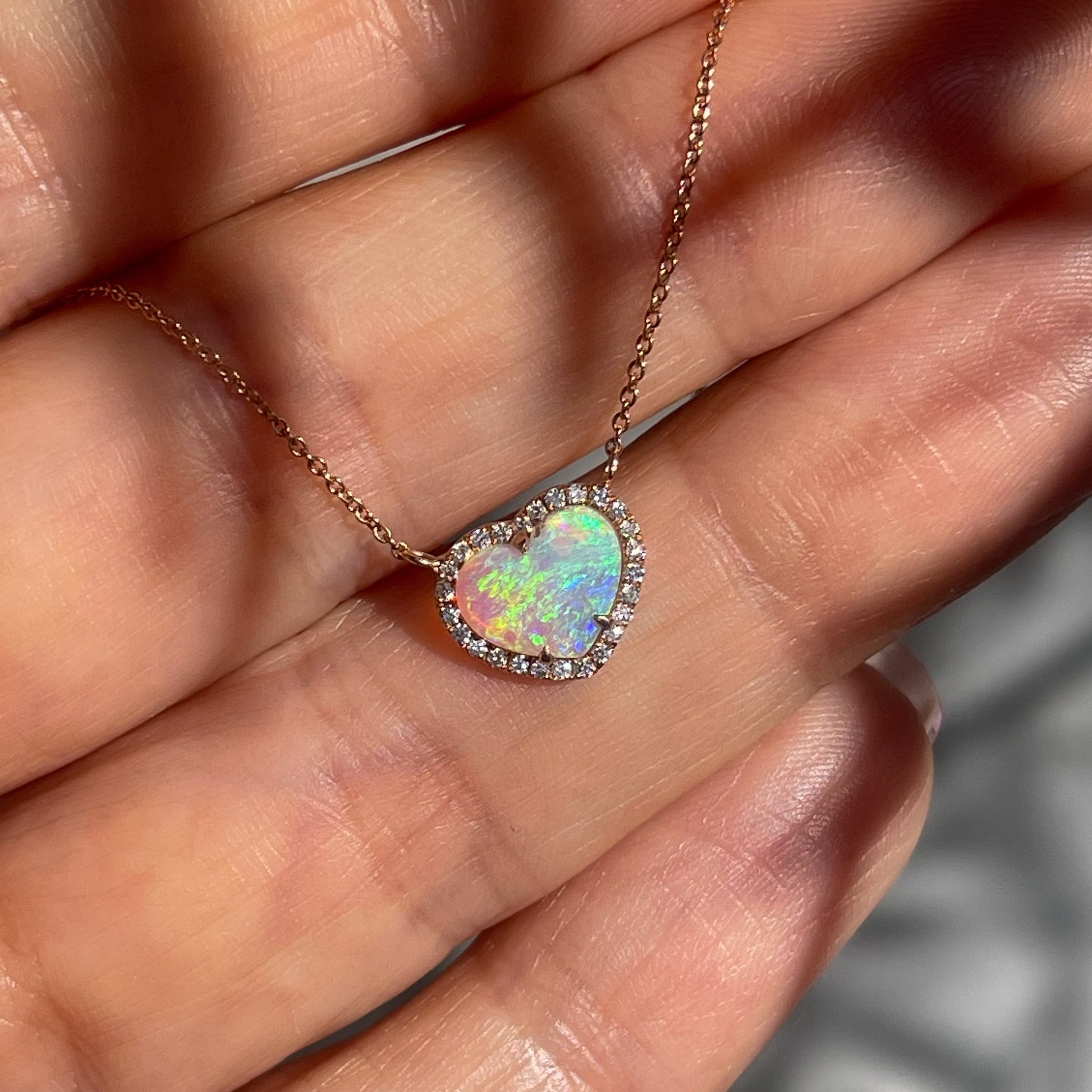 An Opal Heart Necklace by NIXIN Jewelry rests upon a hand  and sparkles in the sun. The natural opal necklace is made of 14k rose gold.
