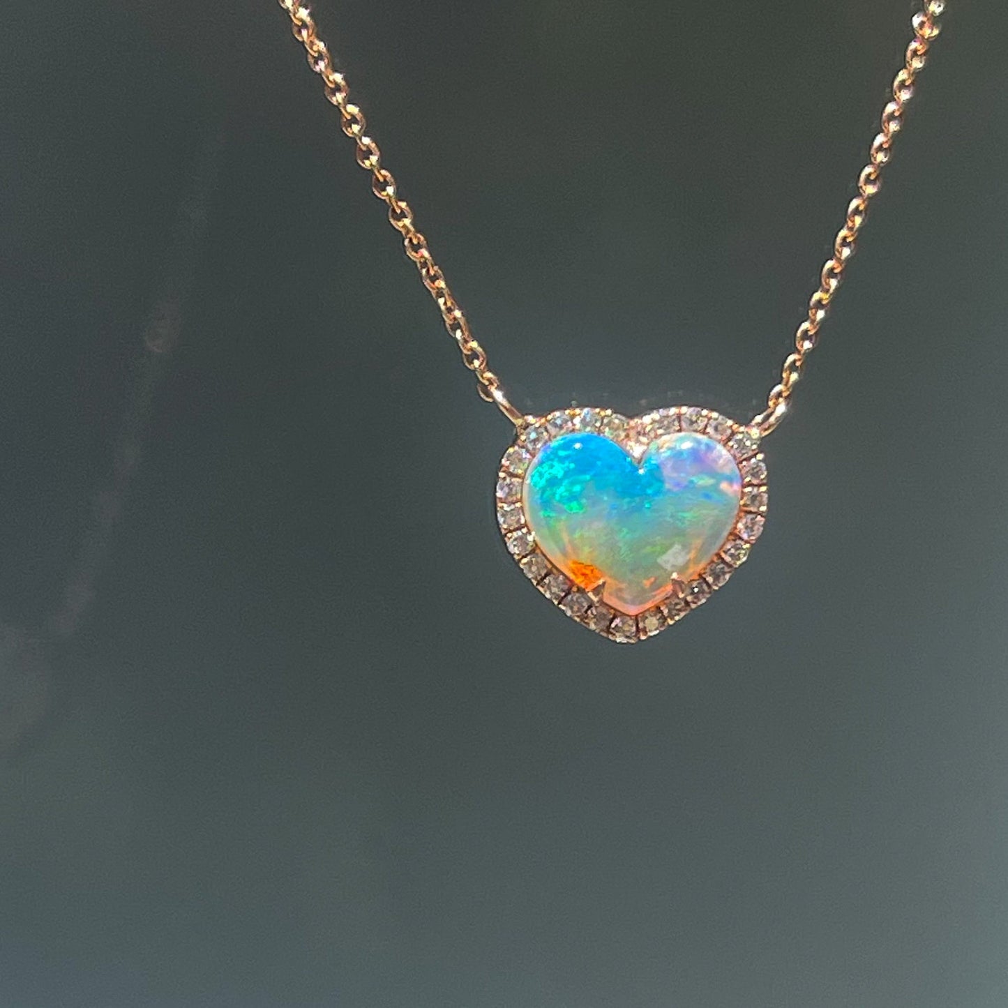 An Opal Heart Necklace by NIXIN Jewelry hangs in front of a frosted glass door. The opal pendant is made in 14k rose gold.