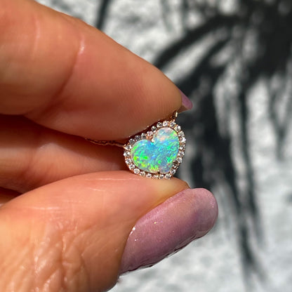 An Opal Heart Necklace by NIXIN Jewelry held up in front of a grey wall. The green opal is secured by prong setting and is surround by a diamond halo.